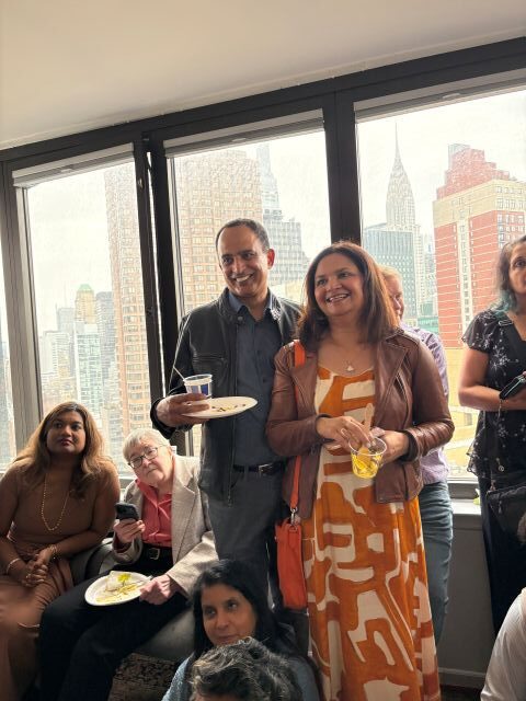 Group of people at a social gathering by large windows with a city skyline outside; a man holds a plate and a woman in an orange dress smiles.