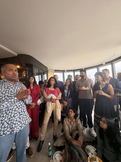 Diverse group listening to a man in a patterned shirt speaking at a casual indoor gathering; some hold plates and drinks.