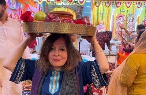 A woman in blue traditional attire balances a large brass platter piled with fruit on her head at a festively decorated wedding hall.